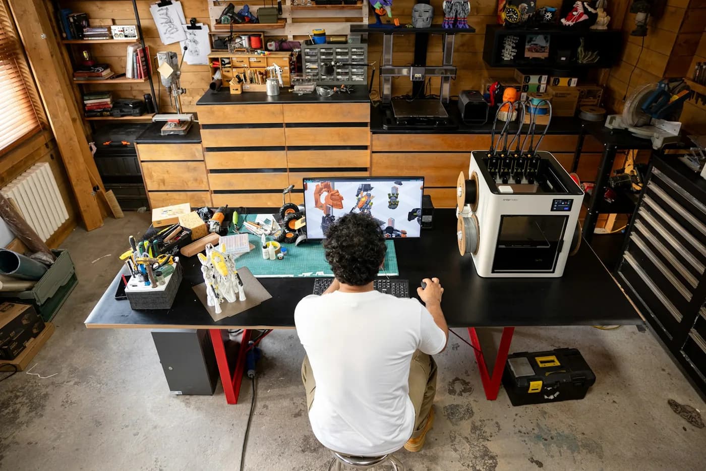 An inventor at a home workbench, modelling a part beside a 3D printer